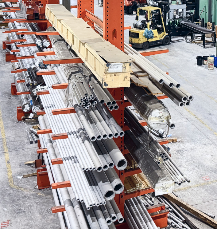 Rows of metal pipes are neatly stacked on red shelving units in a warehouse. A forklift is visible in the background, conveying a busy, industrial atmosphere.